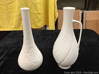 Two white matte textured ceramic vases standing side by side on black cloth with yellow backdrop. One vase displays bulbous base with organic textured pattern, other vase shows tall neck and handle with spiral and circular textured design.