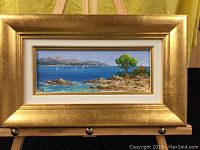 Full view of framed oil painting showing coastal scene with water, rocky shore, tree, and mountains.