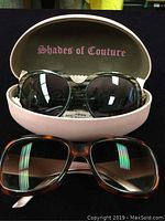 Two pairs of designer sunglasses, one in a pink hard case labeled 'Shades of Couture' and one resting below it with tortoiseshell frames.