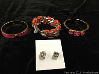 Top view of all four jewelry pieces on a dark background: two Kate Spade bracelets, one Marc Jacobs bracelet with orange woven leather and chain links, and a pair of clear crystal stud earrings on a Kate Spade card.