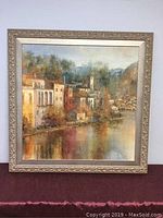 Frontal view of framed acrylic painting showing village by lake with reflections.