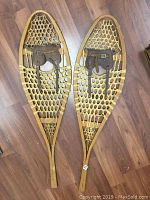 Pair of vintage wooden snowshoes laid flat on wooden floor showing full length with tan woven webbing and brown leather foot bindings.