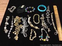 Top-down image of 15 assorted costume jewelry bracelets including bead, charm, cuff, and chain link styles on black background with ruler for scale.