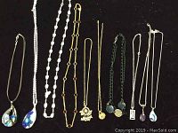 Lot of 10 costume jewelry necklaces displayed on black background, showing variety of pendants and chain styles including glass teardrop and gold-tone chains.