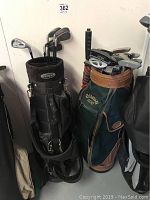 Two golf bags positioned side by side against a wall. The Black Knight golf bag is black with several clubs, the Callaway bag is green with brown accents and a large number of clubs visible.