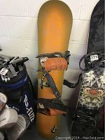 Orange Surf Politics snowboard leaning vertically with black and orange bindings attached. Shows scuff marks and wear on surface.