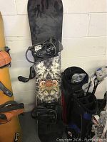 Snowboard leaning against a wall with bindings attached, surrounded by other sports equipment.
