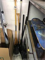 Photo showing the 2 wooden paddles with natural wood finish and 3 metal paddles with black and silver shafts leaning against the wall near a skateboard.