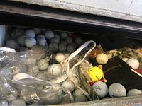 Photo showing a large quantity of white and a few colored used golf balls with some golf accessories.