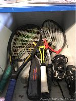 Photo of six tennis rackets with various grip colors and black rackets with logos in a storage bin.
