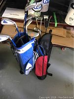 Two youth golf bags placed on the floor with golf clubs inside. One bag is blue and white (Atom), and the other is red and black (Future Tour). Several clubs with head covers and tags visible.