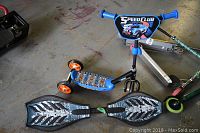 Photo showing the Hot Wheels Speed Club three-wheeled scooter with blue handle grips and front plate, the Ripstik caster board with silver and black design, and a metal framed scooter with green wheels on a concrete floor.