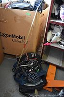 Photo showing three sports bags and hockey equipment stacked beside a shelf and a large cardboard container.