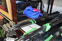 Side view of 12 metal adult golf clubs with various heads bundled together with green ties, placed on a shelf with bags in the background.