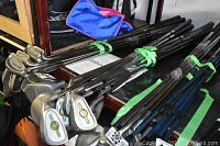 Photo showing multiple golf clubs with metal shafts and variety of golf club heads stacked on a table including irons and drivers. Grips are blue and black.