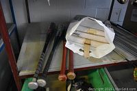 Four bats and a white plastic bag on a shelf; bats include an Easton aluminum, a TPX aluminum, a wooden bat (Cooper), and a Rawlings bat.