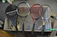 Four tennis rackets with Dunlop Juniors Mid Plus black cover on left side, showing handles and racket heads. Rackets show different brands and colors, resting on flat surface.