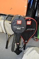 Five tennis rackets with Wilson Profile and Attack Titanium covers leaning against a box in a storage area.