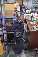 Full view of Sims Odyssey snowboard standing vertically with vibrant purple graphic and attached black bindings.