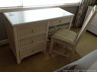 Painted wicker desk with glass cover and three visible drawers under window, paired with chair with striped fabric seat.