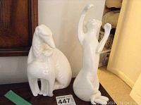 Two white porcelain figurines shown, one with elongated neck and body, another human figure with arms raised.