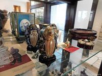 Display of Asian style cloisonné figures, hand-painted egg, bud vase, trinket box, and stone elephant on glass shelf.