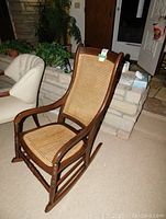 Full view of wooden rocking chair with cane seat and cane backrest in a carpeted room, visible part of an upholstered chair nearby.