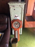 Front view of the distressed green painted wood cabinet, Caravelle wall clock leaning against it, and small metal waste paper bin in front