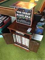 Wooden record cabinet with two doors below and an open shelf space, some records standing inside cabinet, 'The Great Band Era' vinyl displayed on top.