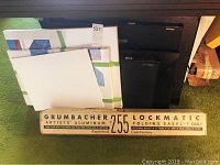 Full view of the Grumbacher 255 Lockmatic aluminum easel box alongside several blank white canvases and black art portfolios stacked on a green carpet.