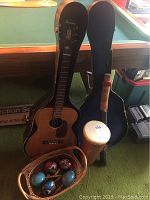 Harmony acoustic guitar with its hard case, small wooden drum with strap, basket containing maracas and mouth harp resting on a green carpet in front of a pool table.