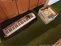 Full view of the Midiman Radium 61 keyboard on green carpet next to a box filled with assorted music books.