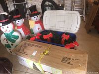 Photo showing three lighted snowmen figures next to a box containing a 7.5 ft artificial Christmas tree, and a plastic bin with wreaths decorated with red bows.