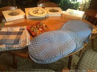 Photo of a round wooden dining table with placemats, napkins, and boxed collector plates on top, showing the fabric placemats and table surface.