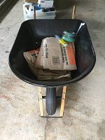 Front view of black wheelbarrow with wooden handles and tire. Inside wheelbarrow are folded paper yard waste bags and various trimmer line packaging.