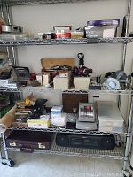 Shelf with mixed office equipment including staplers, cases, adding machine, headphones, camera light, boxes of envelopes and paperclips
