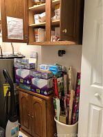 Wood cabinet with open door showing contents including rolls of wrapping paper in a white bucket, and various boxes and containers on shelves.