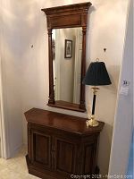 Full view of tall wooden mirror mounted above wooden console with lamp on top.