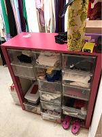 Pink storage cabinet with 15 clear drawers filled with folded clothes and accessories with one drawer open revealing contents.