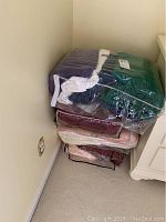 Stack of towels in clear storage bags in various colors including green, purple, black, red, beige, pink, located on carpet floor next to a white nightstand.
