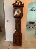 Front full view of wooden grandfather clock with ornate top, glass front, metal lattice lower door, and clock face with Roman numerals