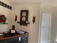Wide angle showing vintage wooden shelf clock mounted on wall between two wooden wall sconces with red candles. TV top below with additional clocks and remotes.