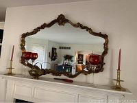 Ornate gold-toned wall mirror hanging above a cabinet, with two brass candle holders each holding a pink taper candle and two ruby red glass candle cups arranged on the cabinet.