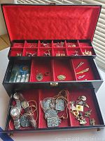 Open jewelry box showing red velvet interior with multiple compartments filled with wristwatches, cufflinks, tie clips, necklaces with pendants, and various pins