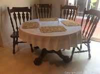 View of round wooden claw foot dining table with white linen tablecloth and four woven placemats, surrounded by four carved wooden chairs.