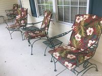 Photo shows two metal rocking chairs with floral cushions, two metal side tables, and two metal chairs with floral cushions arranged on a concrete porch.