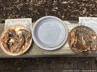 Three collector plates displayed outdoors on a stone surface, including two bronze-colored raised relief plates and one white and gold decorative plate with crest.