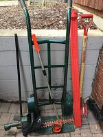 Photo of all items grouped together showing the hand truck, hedge trimmer, level, spade and shingles removing spade leaning against wall