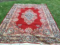 Full view of the 10X8 ft red area carpet displayed on grass, showing the central floral medallion and border patterns, as well as some wrinkles and areas that appear worn.