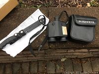 Alcan fishing knife in plastic sheath alongside Bushnell binoculars and binocular case; all items shown with their straps and slight wear visible.